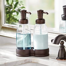 two clear soap pumps with bronze tops and base on a bathroom marble sink top, faucet, mirror, plant